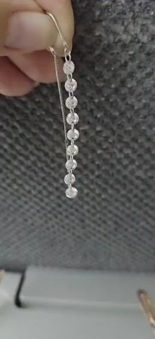 A hand holds a silver earring with a thin chain dangling from it. The chain has multiple small, round, sparkly elements attached in a line. The background is a textured gray surface. The earring appears to be a threader earring, designed to be pulled through the ear piercing.