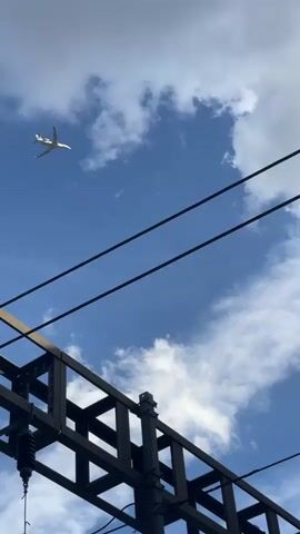 The video captures a clear daytime view of the sky, with a small white airplane flying against a backdrop of scattered white clouds and a vibrant blue sky. The airplane is a prominent subject, positioned in the upper left quadrant of the frame. In the foreground, dark, horizontal wires stretch across the image, likely power lines, creating a layered visual effect. Partially visible at the bottom and right edges of the frame are elements of man-made structures: a dark, angular building on the right and a dark, lattice-like structure on the left, possibly part of a bridge or overhead infrastructure. The lighting suggests it is daytime, with the sun illuminating the clouds and the airplane. The overall impression is one of a fleeting moment of everyday observation, blending the natural elements of the sky with the urban or industrial landscape. There is no visible text.