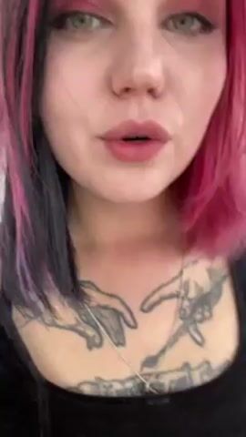 A woman with pink and black hair, red eyeshadow, and a tattoo on her chest is blowing out smoke in front of the camera. She is wearing a black top and has multiple rings on her tattooed hands. The background is slightly blurred, suggesting an indoor setting.