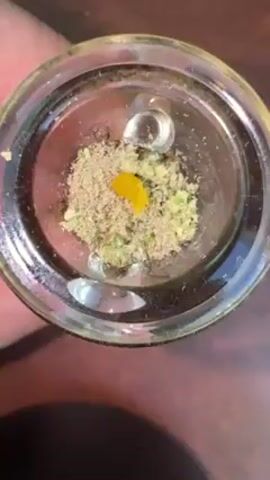 A close-up, top-down shot shows a clear glass bowl containing cannabis flower and a yellow dab of concentrate. The cannabis is a mix of lighter brown, granular material and greener, more leafy pieces.  The yellow concentrate sits slightly off-center. The bowl has two small, clear glass protrusions on opposite sides of the bowl's interior.  The bowl is held by a person whose skin is visible to the left.  The background is a wooden surface.