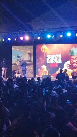 A wide shot shows a crowd of people seated in a dark indoor space, illuminated by stage lighting. A man is onstage, speaking into a microphone. There are two large screens behind him, one showing a video of a man in a suit, and the other displaying a title card that says “Sarkin Dariya True Live”. There is a security guard standing on the left side of the stage. The crowd appears to be an audience at a live event.