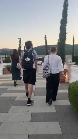 This is a rear view, medium shot video of three people walking away from the camera on a stone pathway. In the foreground, two individuals are walking side-by-side. The person on the left is wearing a black and white patterned short-sleeved shirt, black shorts, and a black backpack. They are facing slightly to the right. The person on the right is wearing a white textured short-sleeved shirt with wide black pants and carrying a black and white striped tote bag over their right shoulder. They are facing slightly to the left. Behind them and further to the left, a third person is walking away from the camera, wearing a dark dress and carrying a beige bag. The pathway has a geometric pattern of light and dark gray rectangular stones. Tall, slender trees in large terracotta pots are visible along the right side of the pathway, flanking a white balustrade. In the background, a body of water is visible, with a rocky cliff face and green vegetation rising behind it. The sky is a pale gradient of blue and yellow, suggesting either dawn or dusk. The overall environment appears to be an outdoor scenic area, possibly a resort or park.