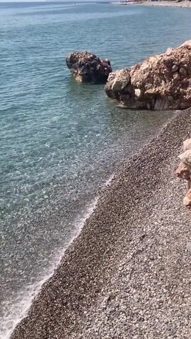 The video shows a tranquil beach scene with clear turquoise water lapping against a pebble shore. Large rocks are partially submerged in the water near the shore, creating a natural barrier. The water is shallow near the edge, revealing the smooth pebbles beneath its surface. In the distance, the horizon meets the calm sea under a bright sky, with a faint outline of a coastline visible. No people or specific objects are prominently featured or interacting. The overall environment is peaceful and natural, indicative of a secluded beach.
