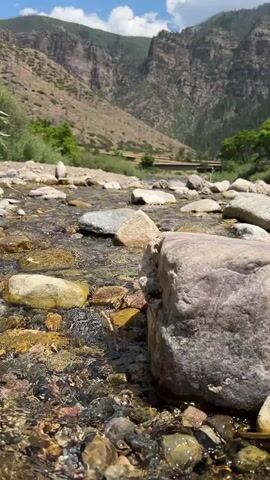 The video shows a close-up shot of a clear stream flowing over a rocky riverbed. The water is shallow and transparent, allowing the colorful stones and pebbles beneath to be clearly visible. Sunlight glints off the water's surface, creating shimmering highlights. In the background, a lush green valley unfolds, with steep, rocky mountains rising dramatically on either side. The sky above is a bright blue, dotted with fluffy white clouds. The overall impression is one of a serene and picturesque natural landscape, untouched by human development.