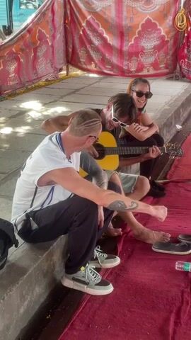 Three individuals are seated together, engaging in a musical moment. A man with a guitar is at the center, playing barefoot while clad in shorts and sunglasses. A woman to his right, also wearing sunglasses, is seated and smiling. To the left, another man sits on a concrete ledge, his attention directed toward the guitarist. This man is sporting a white and blue jersey, dark pants, and sneakers. All three are on a red carpet beneath a canopy made of patterned fabrics. A clear plastic water bottle lies on the carpet.