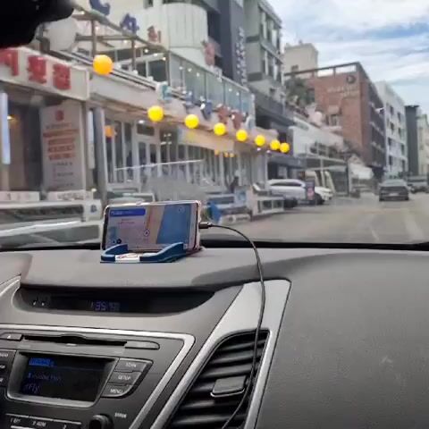 The video shows a view from inside a car as it drives down a street. The car's dashboard is visible in the foreground, with a smartphone mounted on a holder displaying a navigation app. The street is lined with buildings, many of which appear to be shops or restaurants, with signs and awnings. There are other cars on the street, some parked and some moving. The sky is visible above, with clouds. The video appears to be filmed in a city or town with commercial activity.