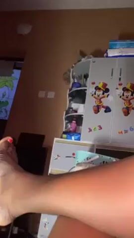 A close-up, static shot shows a person's lower leg and foot, which is angled with the toes pointing upward. The big toe is painted with red nail polish. The leg appears to be resting on a surface, with the background consisting of a wall painted a warm color, along with children's furniture, including a small white cabinet decorated with Mickey Mouse figures and numbers. Part of a window or framed picture featuring a green leafy design is visible in the left background.