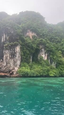 A boat is moving through clear, turquoise water past a steep, jungle-covered cliff face. The cliff is made of rock with some lighter colored sections and is covered in dense green foliage. The water is calm with some ripples and appears shallow near the shore. In the distance, other boats are visible. The sky is overcast.