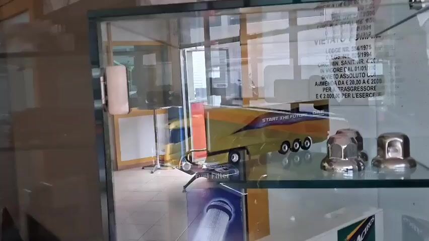The video captures a close-up view of a glass display cabinet, panning downwards and slightly to the left. The setting appears to be an indoor commercial space, possibly a truck dealership or parts store, in Savona, Italy, during daytime.

The top shelf of the cabinet features several miniature truck models. A prominent yellow and black truck model, possibly a DAF, is visible with the text "START THE FUTURE DAF" on its side. Behind this model, a sign is displayed with Italian legal text, including "LEGGE NR. 38/1975", "D. LGS. NR. 26/1994", "CIR. MIN. SAN. NR. 4/2000", "IN VIGORE DAL 01/01/2007", "DIVIETO AL SOLUTO [sic]", "AMMENDA DA € 20,00 A € 200,00 PER IL TRASGRESSORE E € 2.000,00 PER L'ESERCENTE". Next to the yellow truck are several metallic objects resembling lug nuts.

As the camera pans down, a box labeled "Fuel Filter" is visible, likely an actual truck part or a model of its packaging. Below this, another shelf displays more miniature truck models, including a blue and white truck with a trailer and a white truck that appears to have a Mercedes-Benz logo. The reflection in the glass shows a tiled floor and parts of the room's interior. The bottom shelf of the cabinet appears mostly empty, with only a few small items scattered.