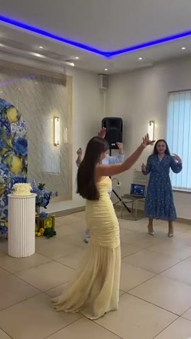 Four women are dancing in a banquet hall. The woman in the foreground is wearing a yellow, strapless gown. Another woman, wearing a blue blazer and jeans, is to the left of the first woman. A third woman, in a blue polka-dot dress, is to the right, and another woman in white is clapping behind her. In the background, there's a speaker, a table with a laptop, and window blinds. The room is decorated with blue lighting accents, a decorative mirror, and flower arrangements.