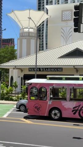 A pink trolley bus is driving down a city street. The bus has "Hi Bus" and "Hawaii's Bus Honolulu" written on its side. A silver car is parked on the left side of the street. Behind the bus and car is a building with a sign that reads "Men's Wearhouse". There is a tall building in the background. The sky is blue and there are some clouds. The scene appears to be in a tropical climate.