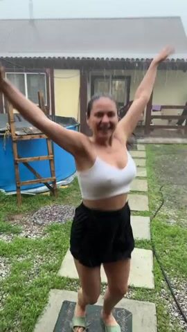 A woman in a white tank top and black shorts is dancing in the rain. She is in a backyard with a house in the background. There is a blue tarp and a wooden structure to her left. She is smiling and her arms are raised in the air. She is wearing flip-flops.