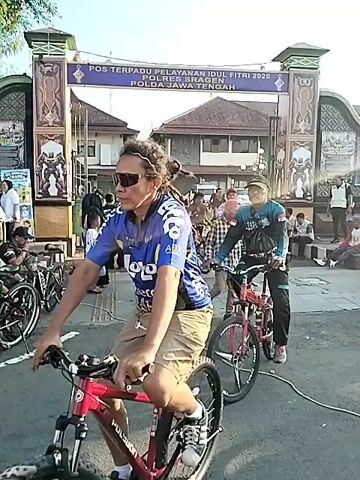 A low-angle, eye-level, wide shot captures a crowd of people participating in a bike ride on a clear, sunny day. The camera pans from left to right, following the cyclists as they ride in a single file line. The cyclists are diverse in age, gender, and attire, with some wearing casual clothes and others sporting cycling gear. The background features a large, ornate archway with text on it, a building behind it, and trees to the left. The archway has text, some in red, that reads "POS TERPADU PELAYANAN IDUL FITRI 2020 POLRES BRAGEN POLDA JAWA TENGAH". The ground is paved, and there are some parked motorcycles visible on the left side of the frame. The overall atmosphere appears to be lively and communal.