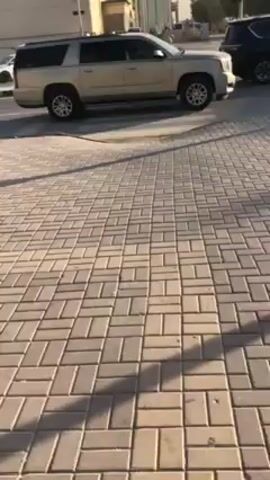 The camera is held at ground level and shows a sidewalk composed of light-tan rectangular paving stones. In the background is a road with moving vehicles including cars and SUVs, of various colors including gold, white and silver. A pedestrian lamp post is seen at the bottom left of the frame. The video appears to be recorded during daylight, casting shadows on the paving stones.