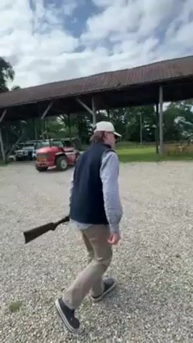 Okay, here's a description of the video:

The video shows a person wearing a light pink collared shirt, a navy vest, and sunglasses, walking across a gravelly area towards a building. They hold the camera in front of them, capturing a first-person perspective. Initially, the video shows a man in a baseball cap carrying a rifle walking away from the camera. The setting appears to be a farm or rural area, with various buildings, including sheds and a larger brick structure.  There are pallets and equipment scattered around the area. The sky is partly cloudy.