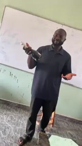 A dark-skinned man stands in a classroom with a bottle of alcohol, possibly champagne or wine. He is wearing a black polo shirt, black pants, and sandals. He is smiling and gesturing with the bottle. There is a whiteboard behind him and a wooden chair to his right. The classroom floor has a speckled tile pattern. The walls are painted in a light green color. There is an electrical outlet on the wall behind him. He appears to be presenting or celebrating something.