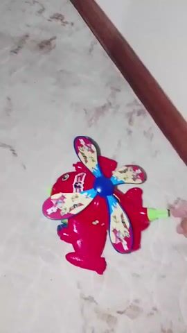 A hand is seen operating a red toy helicopter. The helicopter has a blue propeller with colorful designs. The propeller spins rapidly as the hand pushes and pulls a green lever attached to the back of the toy. The toy is on a light-colored, speckled surface. A dark, wooden object is visible in the upper right corner of the frame. The video is short and shows repetitive action of the toy being operated.