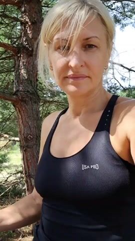 A blonde woman in a black tank top is looking at the camera. She is standing next to a tree. The background shows a forest with green trees and grass.