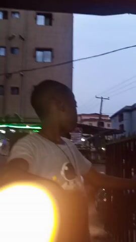 The video shows a child in a gray shirt with an astronaut design smiling at the camera. The child briefly looks off-screen. The camera then pans away, showing a person in a green and yellow patterned shirt behind a metal gate. The camera movement is erratic and shaky. A building is visible in the background.