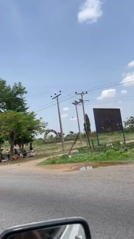A moving vehicle's perspective captures a roadside scene. Initially, several individuals are visible near a green car and a motorcycle, positioned under the shade of trees. The background features an arch-like structure and power lines. The camera then shifts, showing a road lined with vegetation, punctuated by more power lines. Eventually, the view transitions to the interior of the vehicle, with a glimpse of other cars on the road ahead.