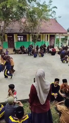 A group of children are gathered in an outdoor courtyard. In the center of the courtyard, several children are engaged in a physical activity, possibly a game or a performance, involving crouching, jumping, and possibly some form of mock combat. Other children are sitting and observing the activity in the foreground and background. The environment appears to be a schoolyard, with buildings visible behind the children, characterized by visible windows, doors, and walls. The ground is a paved surface, with some grassy areas and dirt on the edges. The lighting suggests it is daytime.