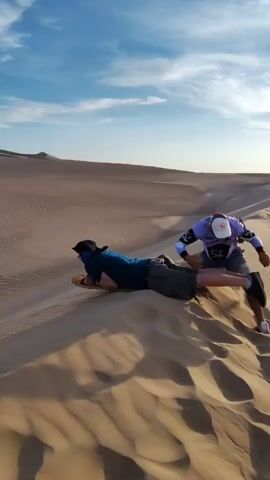 A person is sandboarding in a desert. Another person appears to be helping them get started. The person sandboarding is lying on their stomach and riding down a sand dune. Other people are visible at the bottom of the sand dune. The sky is blue with some clouds.