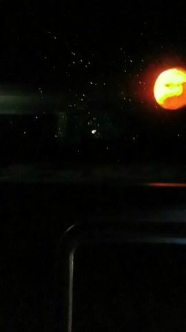 The video is shot from inside a moving car, looking out the windshield at a dark night sky with what appears to be a stylized depiction of a planet or celestial body that is predominantly orange with a green section. There are a few small, faint dots of light, potentially stars, scattered in the sky. As the car moves, the interior dashboard and a portion of the car's frame become more visible. The movement makes the lights appear to streak across the screen. The sounds indicate a conversation is ongoing inside the vehicle.