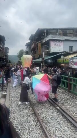 The video shows a crowded street scene, likely in a touristy area, with people standing on railroad tracks. Several individuals are holding large, colorful paper lanterns, preparing to release them. The lanterns are rectangular prisms, with various colors and writing on them. Some lanterns are already airborne, dotting the cloudy sky. Buildings line both sides of the tracks, with shops and food stalls visible. People are wearing casual clothes and appear to be interacting and taking pictures. The ground is covered in gravel.