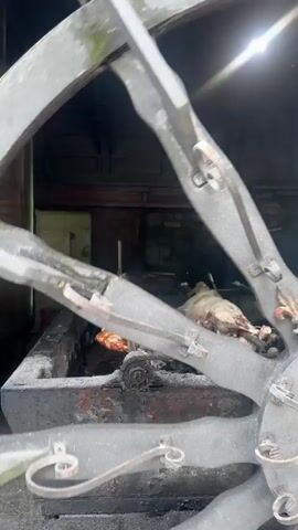A person wearing a red apron is tending to several large cuts of meat roasting over a fire. The meat is on spits and appears to be whole animals. The scene is outdoors, under a wooden roof supported by stone and metal structures. Smoke is visible rising from the roasting meat. There is a large, ornate metal fan-like structure in the foreground, partially obscuring the view of the cooking area. The ground appears to be made of stone and dirt.