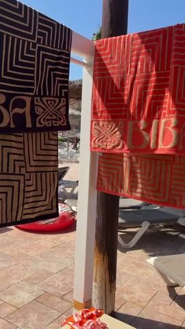 Two patterned towels, one dark and one red, are hanging on a drying rack outdoors. The towels have geometric patterns and the word "BIBA" is visible on both. In the background, lounge chairs and a red inflatable object are scattered around a tiled area. The sky is clear and blue, suggesting it is a sunny day.