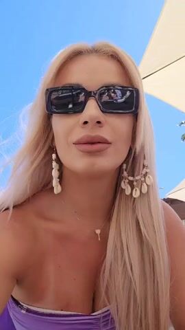 Here is a description of the video:
A woman with long blonde hair is wearing sunglasses and a purple top. She also has on dangling earrings and a necklace with a key pendant. She smiles at the camera. The sky is blue in the background, and there is a white umbrella and a palm tree visible as well.