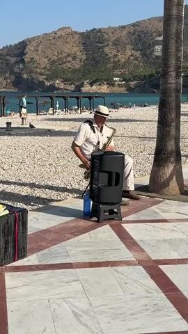 A man in a white shirt and hat plays a saxophone on a sunny day. He is sitting on a stool next to a black speaker, with a blue water bottle at his feet. Behind him, a beach is visible with people relaxing under umbrellas. Mountains rise in the background. The ground is paved with marble tiles in a diamond pattern. To the left, a black case with pink straps is visible. Another man walks by in the foreground, wearing glasses, a dark blue t-shirt, and yellow shorts. The scene appears to be in a seaside town, likely in Spain given the palm tree and mountainous backdrop. The lighting suggests it is late afternoon or early evening. The overall mood is relaxed and pleasant.