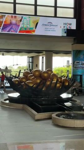 The video shows the interior of a hotel lobby. In the foreground, there is a large, black, metal bowl filled with golden spheres, set on a tiered water feature. Behind the water feature, a large screen displays advertisements and branding for Howard Johnson and "HOW FUN." The screen also shows images of a cityscape and a meal. The background features a large, multi-paned window letting in natural light, and decorative pendant lights made of woven material hang from the ceiling. In the distance, through the lobby and past the window, there are glimpses of trees and sky. Two individuals are seated at a table in the far background, out of focus. The overall environment appears to be a spacious and modern hotel lobby.