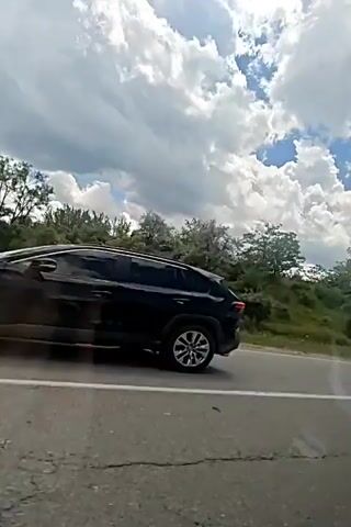 The video shows a roadside view of a green, hilly landscape under a sky with dramatic clouds. A paved road with a white line runs along the bottom of the frame. Trees and bushes are scattered across the hillside. The camera appears to be moving, likely from inside a vehicle, as evidenced by the blurred foreground and the passing of a white car on the road. The overall impression is a scenic drive on a partly cloudy day.
