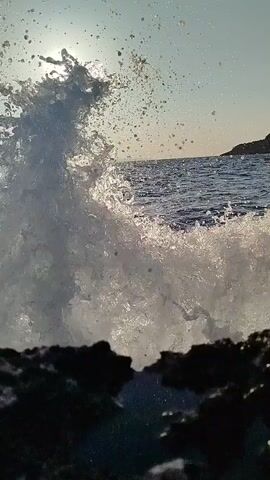 A video captures a wave crashing against rocks by the sea under a bright sun. The sun is visible in the upper left corner of the frame, casting a glare on the water. The waves are choppy and white with foam. There are rocks in the foreground of the video. The overall scene is one of natural beauty and the power of the ocean.