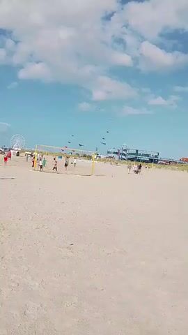 A wide shot of a beach with a bright blue sky and scattered white clouds. In the foreground is a sandy beach. In the middle ground, there is a yellow volleyball net set up with several people playing volleyball. Further in the background, there are more people and buildings, and a Ferris wheel can be seen on the left. In the sky, there are several kites being flown.