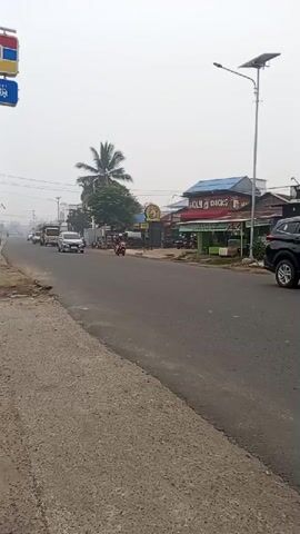 A hazy, overcast sky hangs over a two-lane asphalt road. On the left side of the road, a sidewalk made of cracked concrete leads to a raised dirt area. A sign for "Indomaret" with red, blue, and yellow elements is visible in the upper left corner. Further down the road, several vehicles are moving: a white sedan, a yellow truck, and a motorcycle. The buildings lining the right side of the road appear to be shops, one with a sign that reads "MOLY CHICKS." A solar-powered street lamp stands on the right side of the road. The overall environment suggests a somewhat rural or suburban roadside scene with light traffic.