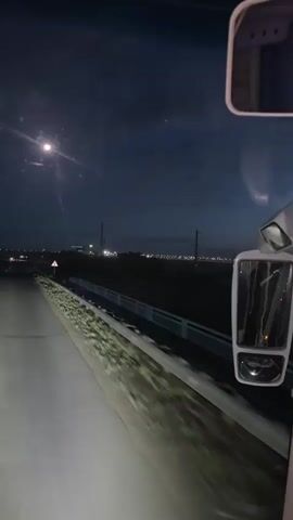 The video captures a nighttime drive through the rural landscape of Kökpekti, Kazakhstan. The perspective is from inside a vehicle, likely a bus or truck, looking forward through the windshield. The road ahead is illuminated by headlights, casting a bright, sharp beam that stretches into the darkness. The sky is a deep, inky blue, with a few scattered lights suggesting a distant town or industrial area. A solitary streetlight illuminates a small section of the road, its light creating a halo effect in the night air.

On either side of the road, the terrain is dark and indistinct, with hints of trees and vegetation. A white-railed barrier runs along the right side of the road, and beyond that, a slope covered in greenery. A triangular warning sign with a red border is visible on the right side of the road, though its specific warning is obscured by the darkness and distance.

The overall mood is one of quiet, solitary travel through the night. The absence of other vehicles on the road, save for distant red lights that could be other vehicles, emphasizes the sense of being on a journey through a sparsely populated area. The bright headlights cutting through the darkness create a feeling of progress and movement, despite the stillness of the surrounding landscape. The time of day is clearly night, given the dark sky and artificial lighting. There are no people visible, and the objects are limited to the road, the vehicle's interior, and the surrounding landscape features. The location cues are limited to the general impression of a rural highway in Kazakhstan.