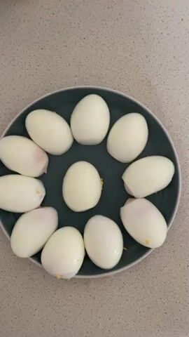 The media file shows a top-down view of a dark grey plate filled with peeled hard-boiled eggs. The eggs are arranged in a circular pattern, with one egg placed in the center and the rest forming a ring around it. The eggs appear to be whole, with no visible signs of being cut or prepared for deviled eggs. The plate is set on a speckled countertop, likely in a kitchen setting. The lighting suggests it is daytime, possibly indoors. There are no people visible in the media file, nor any discernible text. The overall scene is simple and focused on the arrangement of the eggs.