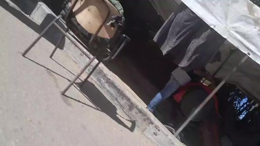 The video shows several green propane tanks lined up on a sandy or dirt ground. In the background, there are tents and a corrugated metal fence. A person wearing a light-colored shirt is visible near the tents. The sky is blue and clear, suggesting it is daytime. The camera perspective is low and moves somewhat erratically, giving a shaky view of the scene.