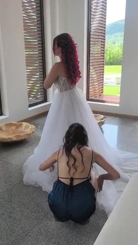 A woman in a white wedding gown with long, dark red hair stands posing in front of a mirror, admiring herself. Another woman with long, dark curly hair, wearing a dark blue, backless dress, crouches on the floor, holding a camera and taking photos of the bride. The room is bright, with large windows featuring wooden blinds and a large, shell-shaped decorative bowl on the floor.