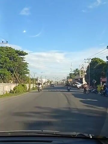 The video shows a street scene, likely in a town or city, from the perspective of a vehicle driving forward. The sky is clear blue with a few wispy clouds. On either side of the road, there are buildings, trees, and utility poles with wires. Several motorcycles are visible, some moving and some parked. People are seen on and around the motorcycles. The road has painted lane markers. The overall impression is a bright, daytime scene in a somewhat developed area.