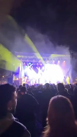 The video shows a live outdoor concert at night. A band is performing on stage with bright purple, yellow, and white spotlights shining on them.  A crowd is gathered in front of the stage, their faces and features obscured by darkness and the backs of other people's heads.  A woman in the band, wearing a hat and a blue shirt, sings into a microphone while holding a trumpet.  She gestures with her arms as she performs.  The stage is surrounded by scaffolding and lighting equipment.  There is some fog or smoke in the air.