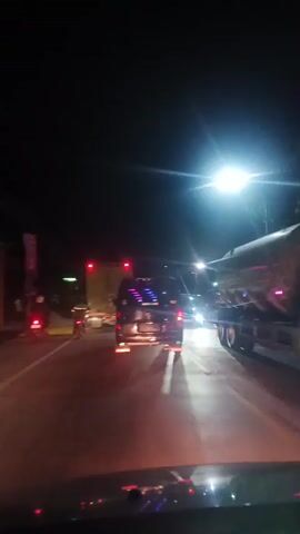 The video shows a nighttime view from inside a vehicle traveling on a road with other vehicles. The scene is illuminated by streetlights and the headlights of other cars. Several vehicles are visible, including a truck with its taillights on, a dark-colored van or SUV with its headlights on, and two motorcycles. The road appears to be paved and has lane markings. The overall atmosphere is dark with bright lights reflecting off the wet road. The video is shaky, suggesting it was recorded while the vehicle was in motion.