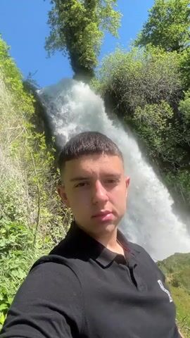 A young man stands in front of a large waterfall on a sunny day. He is wearing a black polo shirt and has short brown hair. He looks into the camera, then turns to his side to look at the waterfall. The waterfall is surrounded by green trees and vegetation, and the sky is blue.