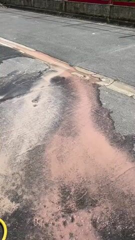 The video shows a street with a pink liquid pooling and spreading across the asphalt. The liquid appears to be flowing from the curb area onto the street. The overall scene suggests a spill or leakage of a pink-colored substance. A yellow hose is partially visible at the bottom left corner of the frame. A pink railing can be seen in the background.