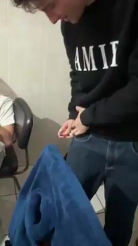 Here is a description of the video:

The video shows a young man standing in what appears to be a bathroom. He is wearing a black sweatshirt with "AMIRI" printed on it, blue jeans, and a belt with a large buckle. He is holding something small and red in his hands, possibly a lighter. He reaches down into his pocket, briefly pulls the object out, and then reacts suddenly, startled or in pain, jerking his leg and yelling. There is a chair with a person sitting in it to the left of the frame, and a blue towel is draped over something in the foreground. The room is tiled with white tiles.