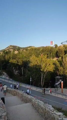 The video pans across a modern amusement park or recreational area situated on a hillside overlooking the sea. In the foreground, a paved pathway is marked with colorful circles. People are walking along this path. Behind them, there are several buildings with white and red facades, some with outdoor seating areas. Trees and rocky terrain are visible on the hillside. In the distance, a roller coaster track is situated on a higher section of the hill, and a flag is flying from a pole. The sky is clear and blue, indicating daytime.