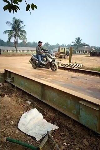 A person on a motorcycle is on a scale. The person is wearing a dark shirt and jeans. The motorcycle is silver. The scale is made of metal. In the background, there are buildings, trees, and a clear sky. The person is riding the motorcycle forward. The camera is filming from the side.