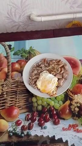 The image displays a close-up of a prepared meal presented on a white, round plate. The plate contains a serving of buckwheat, pieces of cooked meat with sautéed onions, and a fried egg with a visible yellow yolk.

The plate rests on a decorative table mat or tablecloth that features a vibrant still life of various fruits and berries. Visible fruits and berries include green grapes, red cherries, red currants, blueberries, red apples, yellow pears, and a slice of watermelon. Some of the fruit, specifically apples and a mix of grapes, pears, and currants, are depicted in woven baskets on the mat. The mat itself has a patterned white background, possibly resembling lace or a floral design.

In the background, a light-colored wall with a subtle textured or patterned wallpaper is visible. A white pipe runs horizontally across the upper part of the frame, below the wall. To the upper right, a coiled yellow electrical wire is present. The meal appears to be set on a dark-colored wooden table or shelf.

The scene depicts a simple, home-cooked meal in a domestic setting within Жамчужненскі сельскі Савет, Belarus. No individuals are present, and no specific activity is occurring other than the static presentation of the food. There is no visible text in the image.